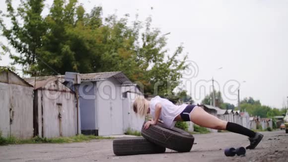 运动年轻的金发女人穿短裤在轮胎俯卧撑的帮助下进行各种力量练习视频的预览图