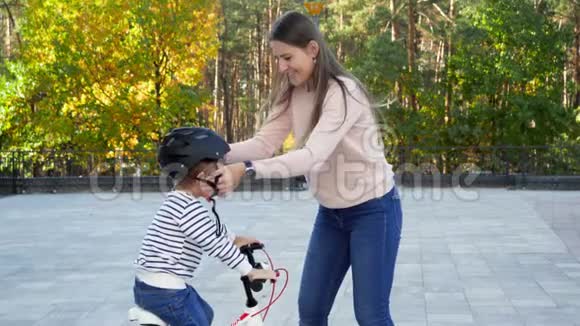 在公园里一位年轻有爱心的母亲在她的小儿子身上戴上并系上了保护自行车头盔视频的预览图