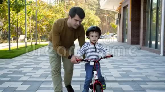 4K镜头快乐的小男孩戴着防护头盔和他的年轻父亲在城市街道上骑自行车视频的预览图