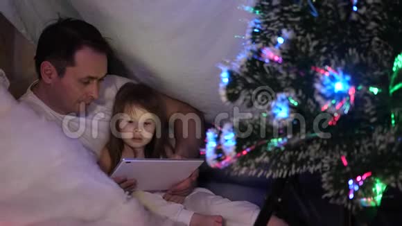 父亲和女儿在圣诞节晚上在平板电脑上玩和看动画片在孩子们的房间里有一个帐篷里面有一个帐篷视频的预览图