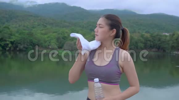 年轻健康的亚洲女人在一个带湖的户外自然公园晨跑时停下来喝水擦汗视频的预览图