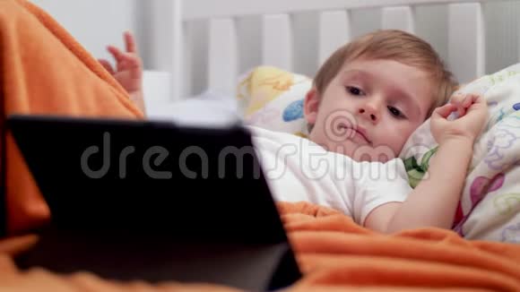 4k特写视频一个可爱的微笑男孩躺在床上在平板电脑上看动画片时微笑视频的预览图