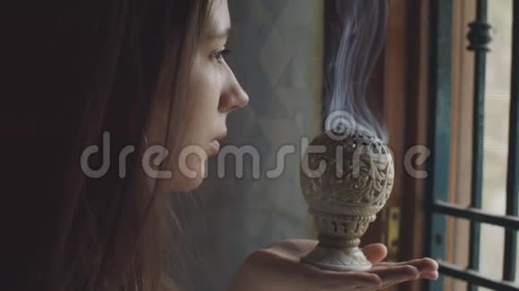 沉思的女人在室内黑暗的背景下通过燃烧香炉冥想抱着印度香沉思的女孩视频的预览图
