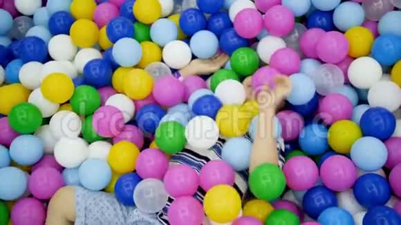 4k视频快乐地笑着蹒跚学步的男孩躺在游泳池里在操场上购物时玩着五颜六色的塑料球视频的预览图