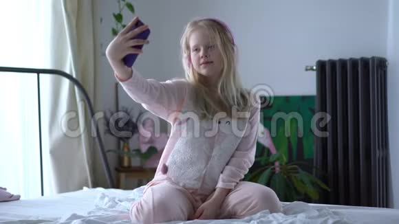 现代生活中的Z一代十几岁的女孩穿着睡衣戴着耳机在床上听着音乐视频的预览图