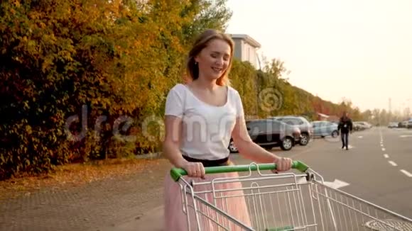 4K慢动作视频美丽的微笑女孩穿着粉红色裙子在停车场和购物中心与购物车散步视频的预览图