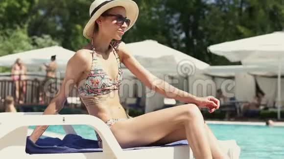 适合年轻的比基尼女士戴帽子坐在游泳池边缘的桌椅和日光浴视频的预览图