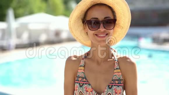 穿着比基尼戴着帽子和太阳镜的漂亮年轻女孩在游泳池的背景下快乐地笑着视频的预览图