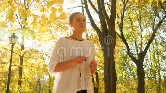 美丽的微笑女孩戴着耳机在美丽的秋天公园慢跑的4k慢镜头视频的预览图