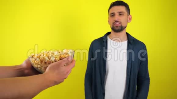 一个穿着蓝色衬衫和白色T恤的年轻人拒绝了一个带有爆米花和垃圾食品的玻璃盘子的建议视频的预览图