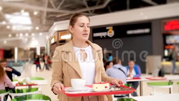 4k视频年轻微笑的女人拿着托盘拿着食物在购物中心的食物场里找个地方坐着吃视频的预览图