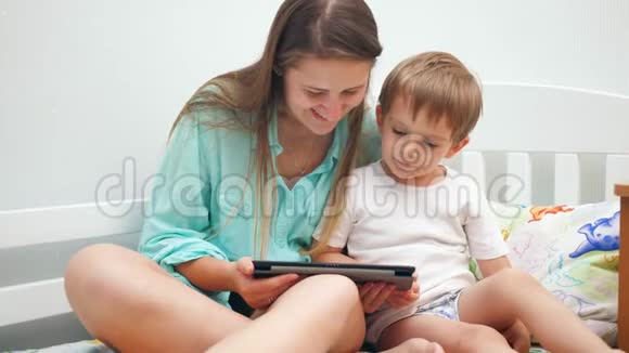 年轻母亲和小儿子坐在床上使用平板电脑的4k视频视频的预览图