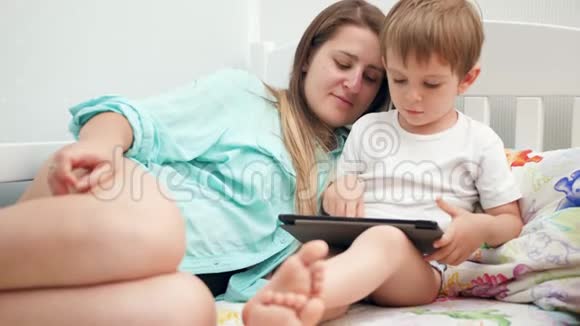 4k视频小学步的孩子年轻妈妈穿着睡衣躺在床上在平板电脑上玩游戏视频的预览图