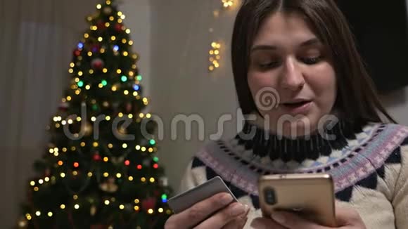 圣诞树附近的一个年轻女孩带着一部电话用信用卡支付新年礼物新年视频的预览图