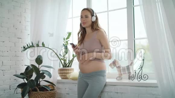 微笑的准妈妈在耳机里听音乐在家跳舞的孕妇视频的预览图