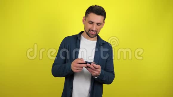 一个穿着蓝色衬衫和白色T恤的年轻人热衷于在智能手机上玩游戏视频的预览图