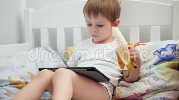 4k视频可爱的小男孩躺在床上一边吃香蕉一边用数码平板电脑视频的预览图