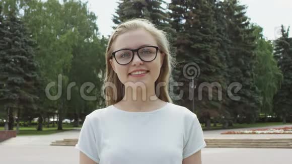 戴着眼镜的漂亮的金发女郎开心地笑着在镜头前看着站在公园广场上的镜头视频的预览图
