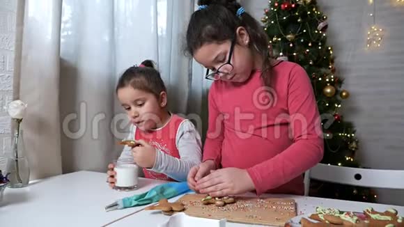 圣诞树背景上的两个小女孩小姐姐一个女孩喝牛奶和姜饼饼干视频的预览图