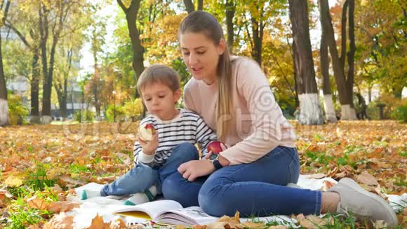 年轻母亲在秋天公园野餐时给她的小儿子看书和吃苹果的4K镜头视频的预览图