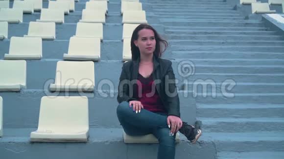 一个穿着自行车夹克的漂亮而热情的女孩独自坐在一个空荡荡的体育场里女孩头发在风中发展视频的预览图