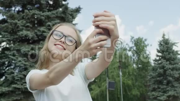 戴着眼镜的漂亮年轻女子在针叶树和蓝树面前的自拍照上笑容灿烂而欢乐视频的预览图