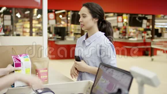漂亮的微笑女孩在超市的收银台等待收银员把她的东西打包成纸视频的预览图