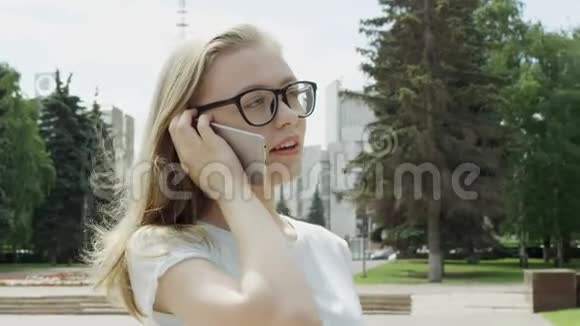 阳光明媚的一天在手机旁的公园里一位戴眼镜的漂亮年轻女性在户外接听电话视频的预览图