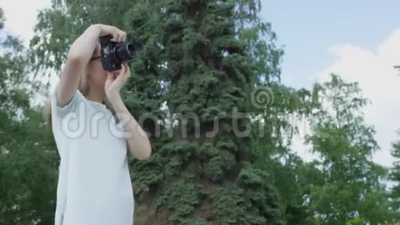 在夏季阳光明媚的日子里金发年轻女性穿着白色衣服在公园里用相机拍摄一系列照片视频的预览图