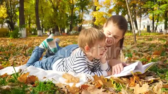 在秋天公园小男孩躺在草地上和黄叶上带着他一起看故事书的4K镜头视频的预览图