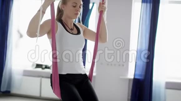 健康的女人在瑜伽课上倒吊在瑜伽吊床上在室内伸展双腿用手握住它们视频的预览图