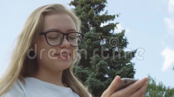漂亮的戴眼镜的金发女人蓝眼睛专注地发着短信脸上带着微笑站在蓝天上视频的预览图