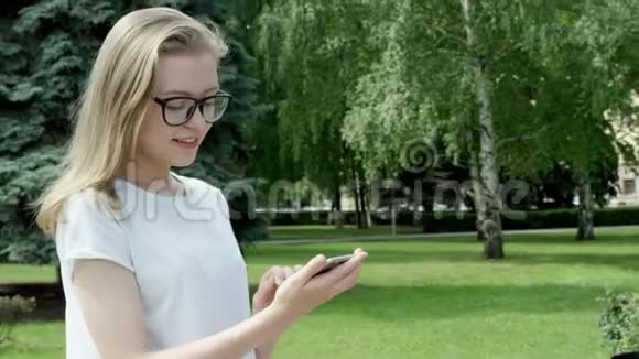 迷人的年轻女性在阳光明媚的日子里全神贯注地使用智能手机聊天金发女孩站在公园前面视频的预览图