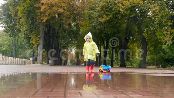 雨天公园里一个蹒跚学步的小男孩带着玩具车在绳子上穿过大水坑的慢镜头视频的预览图