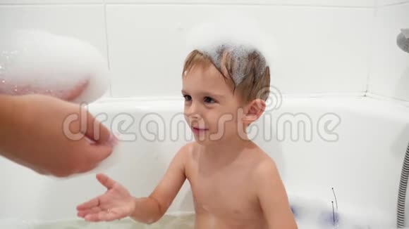 年轻母亲在洗澡时给小儿子洗澡和玩肥皂泡沫的慢动作视频视频的预览图