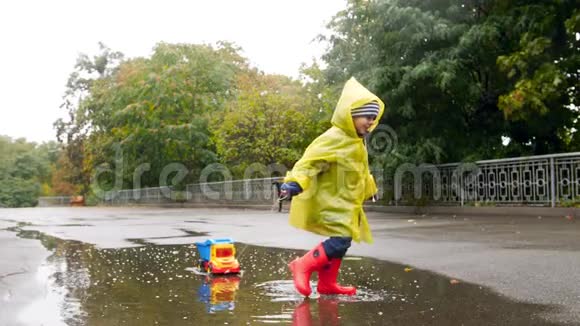 一个穿着橡胶靴的快乐小男孩在玩具卡车的大水坑里奔跑玩耍的视频视频的预览图