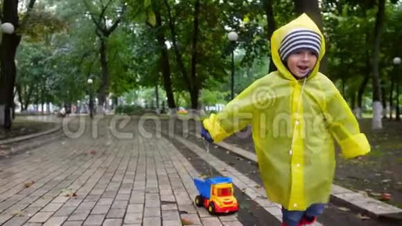 4k视频穿着雨衣在潮湿的秋天公园里奔跑用绳子拉玩具卡车的快乐欢笑小男孩视频的预览图