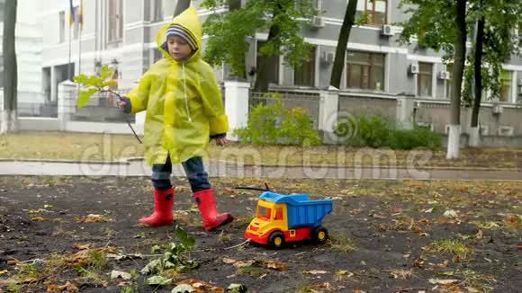 4k小男孩穿着橡胶靴和雨衣用绳子拉玩具卡车收集秋天落叶视频的预览图