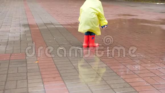 雨后穿着红色橡胶靴和黄色雨衣的小男孩在大水坑里跳跃的慢动作视频视频的预览图