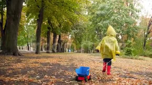 4k视频穿雨衣的小男孩在公园的秋天落叶上散步用绳子拉玩具卡车视频的预览图