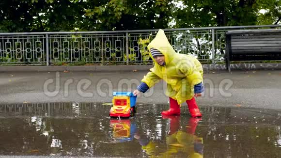 雨后穿着橡胶靴和雨衣的小学步男孩在大水坑里与玩具卡车相撞的慢动作视频视频的预览图