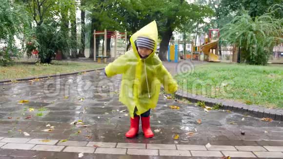 4k慢动作视频穿着雨衣和惠灵顿靴子的快乐小男孩在水坑里跳在水中泼水视频的预览图