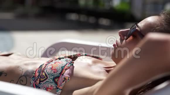美丽的白种人比基尼女孩戴着帽子坐在游泳池边缘的桌椅和日光浴视频的预览图