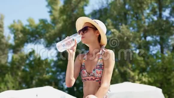美丽的比基尼女郎戴着帽子戴着太阳镜从绿树和蓝天上的塑料瓶中喝水视频的预览图
