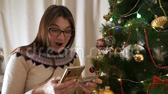 圣诞树附近的一个年轻女孩带着一部电话用信用卡支付新年礼物新年视频的预览图