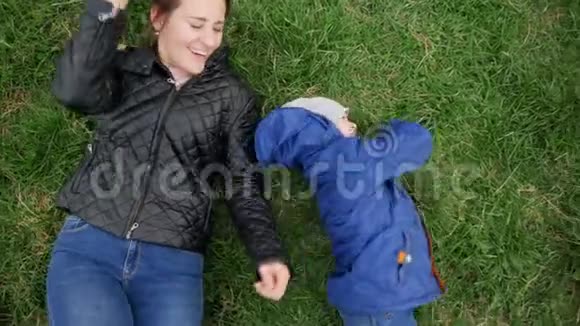 4k视频从高点快乐的笑着年轻妈妈和小儿子躺在公园的绿草上视频的预览图