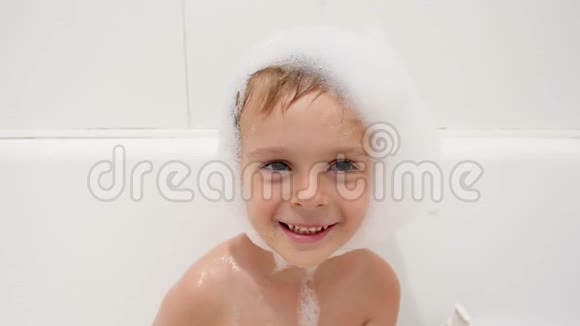 特写4k镜头的快乐微笑的小男孩头上和脸上有很多肥皂泡沫坐着洗澡视频的预览图