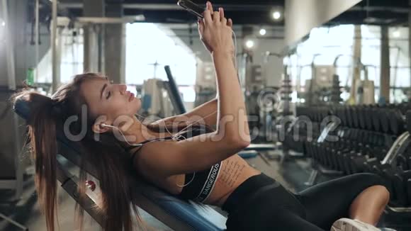 年轻女子带着耳机在健身房锻炼后听音乐微笑迷人的女孩放松和休息视频的预览图