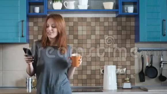 可爱的红发女人在智能手机上打开音乐在舒适的厨房里跳舞视频的预览图