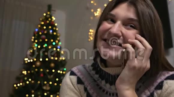 圣诞树附近的女人漂亮的女性使用手机获得新年祝贺年轻女子视频的预览图